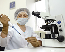 A scientist is placing liquid into a test tube via pipette while sitting in front of a microscope.