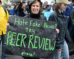 A woman holding a sign that says "Hate fake news? Try peer review... please!"