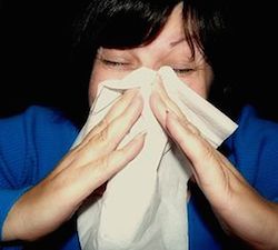 a woman's face close up. Her bangs fall in her face as she pushes her face in to a tissue. She holds the tissue to her face with both hands. She may be sneezing, or just blowing her nose.
