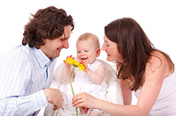 A family, with the dad on the left and mom on the right, sit on either side of their infant child. The child is sitting in a small beige chair. All members of the family wear white clothing, and a white background sits behind them.